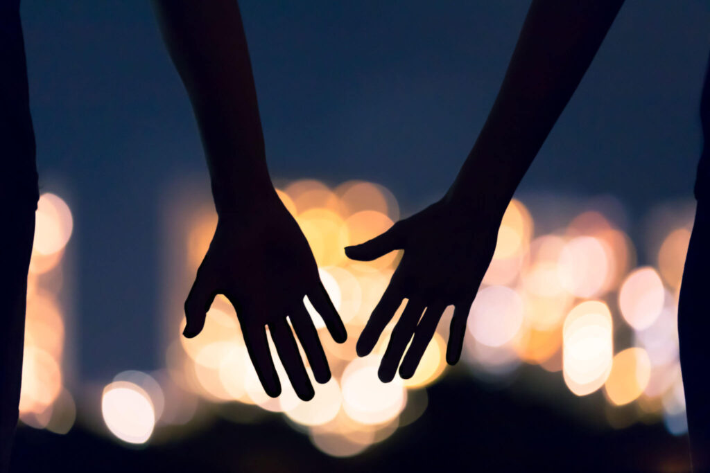 A close up of two hands reaching toward each other against the light of city night lights. This could represent the bonds cultivated by attending multicultural marriage counseling. Learn more about interracial relationships in South Bay, CA, and how a couples therapist in Hermosa Beach, CA can offer support.
